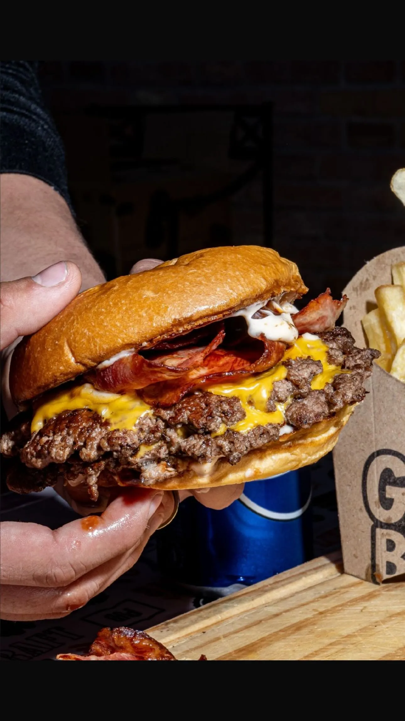 Doble smash con bacon, cheddar y salsa, sostenida en mano junto a papas fritas